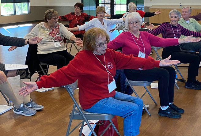 A group of seniors performing stretches while sitting on chairs in a gymnasium.