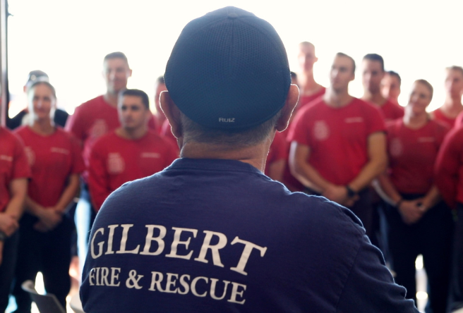 A firefighter in a blue uniform in the foreground with fire recruits in red shirts in the background.