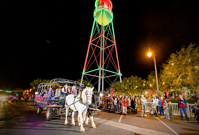 A horse-drawn carriage makes its way through the Gilbert Water Tower Plaza covered in holiday lights. The town's iconic water tower is light red and green in the distance.