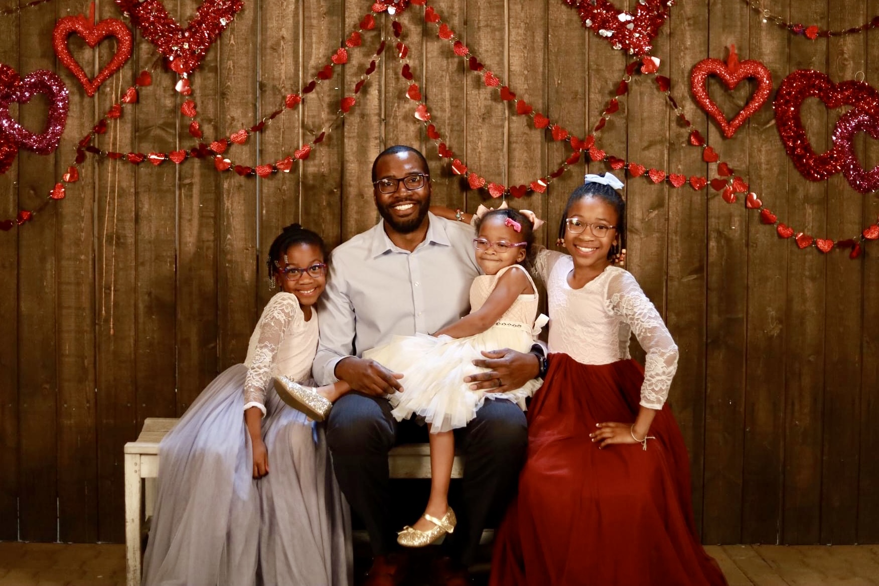 A father poses with his three daughters in front of a wooden backdrop covered in red garland at Gilbert's 2022 Father/Daughter Dance.