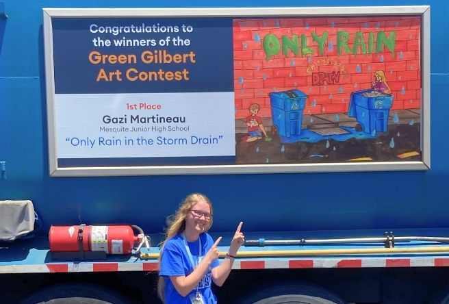 Gazi Martineau poses in front of her winning art design on the side of a Gilbert garbage truck. A sign congratulates her for being one of the winners.