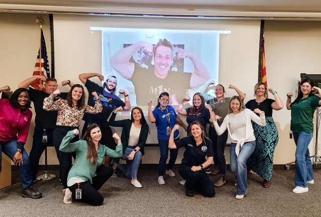 A group of Gilbert employees and a speaker on a project screen all pose for the camera while flexing their muscles.