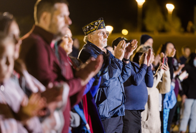 A crowd is seen standing and clapping. A veteran in the crowd is in focus.