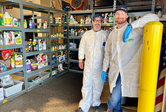 Two male technicians pose for a photo in front of shelves of household hazardous waste items.