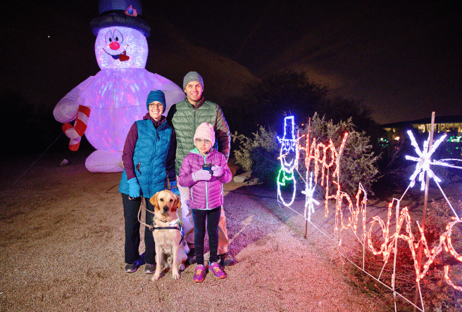 A small family with a mom, dad, child and dog pose in their winter clothes in front of holiday lights at the Riparian.