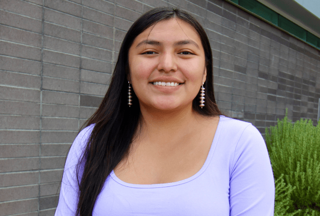 NAMI intern, Octavia Freeland, poses for a photo in front of a building with a green bush. She is smiling and looking directly into the camera.