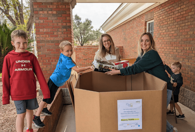 Two women and three young boys stand around a large donation box. The women are holding a donation item above the box.