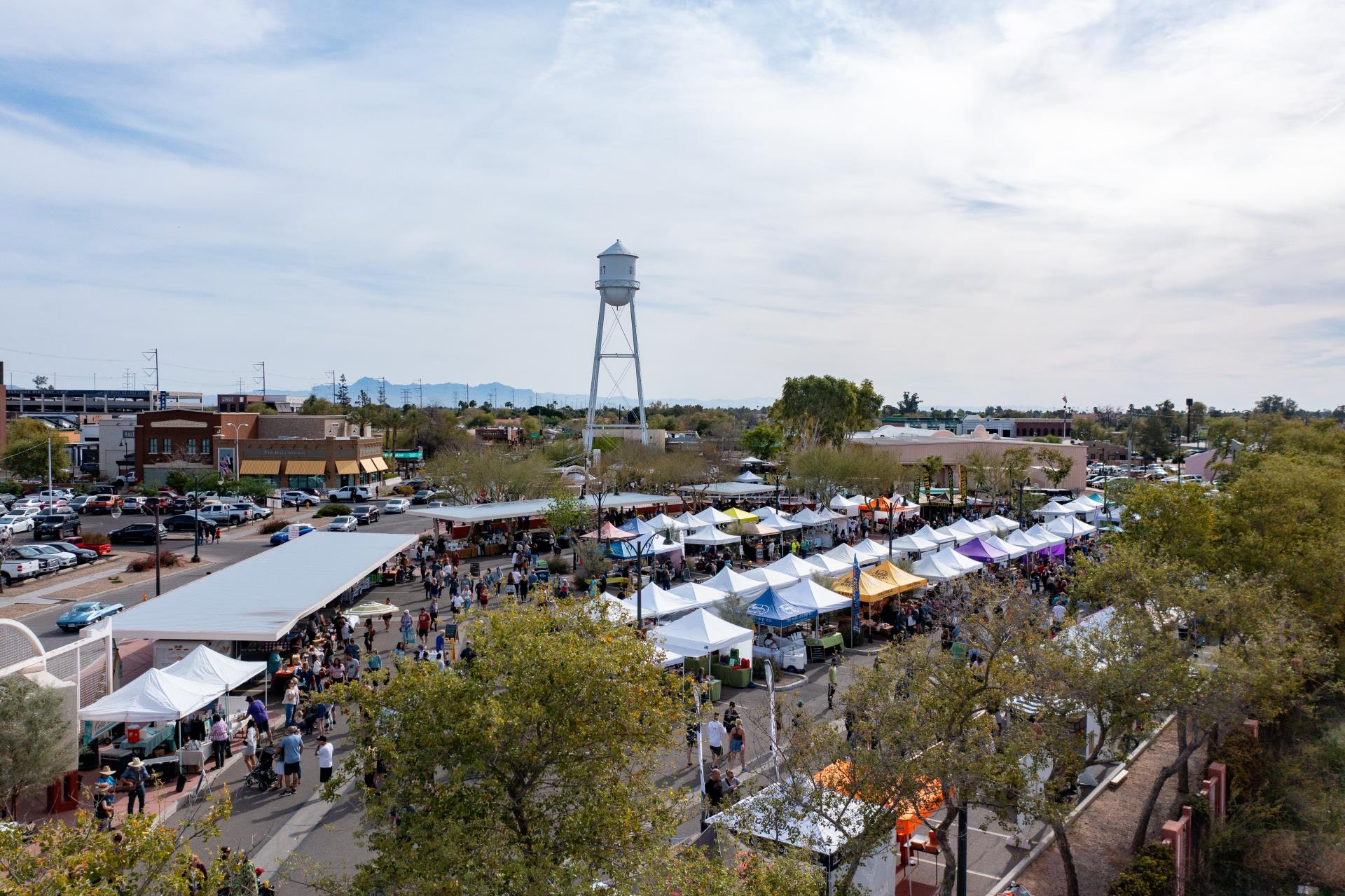 A wide shot photo of the Gilbert Farmers Market with the Water Tower in the background and trees in the foreground. The big blue sky is seen above.