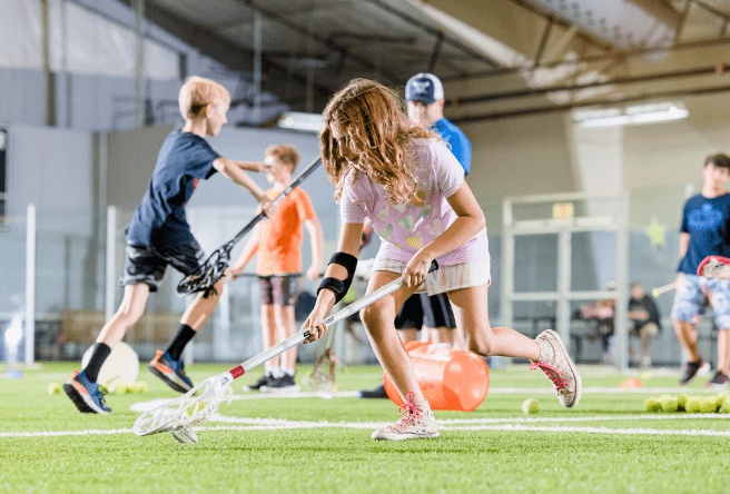 A young girl and young boys are seen playing lacrosse at an indoor field with a male coach wearing a baseball cap in the background.